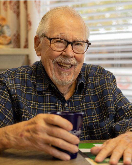 An elderly man in a plaid shirt smiling while holding a coffee mug at a table, symbolizing a comfortable and secure retirement.